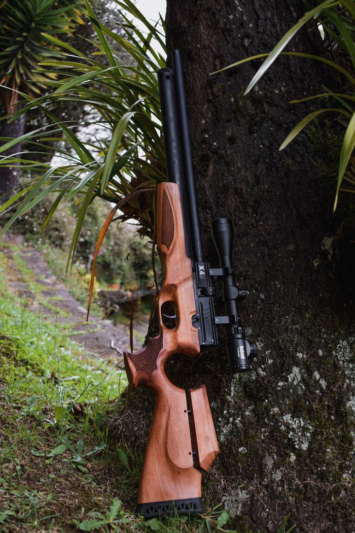 A close-up of a wooden rifle with a scope leaning against a tree trunk amidst green foliage.