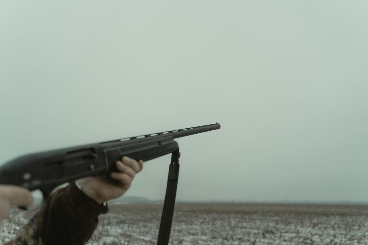 A person aiming a rifle in a vast open countryside setting on a cloudy day.